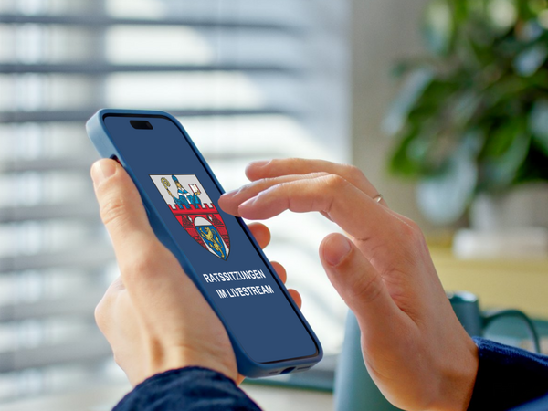 Das auf dem Smartphone abgebildete Wappen gehört zur Stadt Siegen in Nordrhein-Westfalen. Die Stadt Siegen bietet, wie der Text im Bild "RATSSITZUNGEN IM LIVESTREAM" zeigt, die Übertragung ihrer Ratssitzungen als Livestream an.