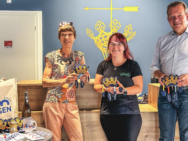 (1)	(v.l.n.r.) Astrid Schneider (Leiterin Kulturabteilung der Stadt Siegen), Sandra Schmidt (Mädelskramladen) und Bürgermeister Steffen Mues präsentieren das Jubiläumsmaskottchen "Niki" in der Touristinformation. (Quelle: Stadtmarketing Siegen GmbH)