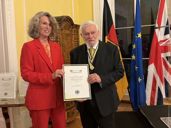 Botschafterin Sabine Baumann (links) überreicht Robert Gettings (rechts) eine Urkunde zu seiner Auszeichnung.