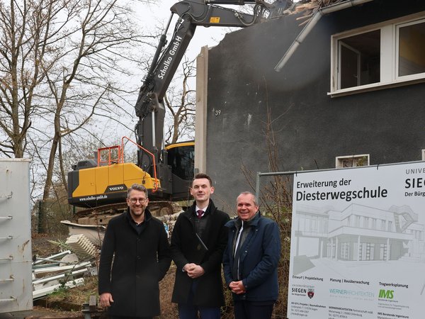 Stadtbaurat Henrik Schumann, Bürgermeister Tristan Vitt und Schuldezernent Andree Schmidt vor dem alten Hausmeistergebäude. Im Hintergrund reißt an Bagger das Gebäude ab.