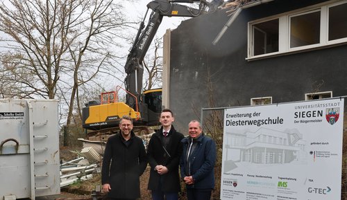 Stadtbaurat Henrik Schumann, Bürgermeister Tristan Vitt und Schuldezernent Andree Schmidt vor dem alten Hausmeistergebäude. Im Hintergrund reißt an Bagger das Gebäude ab.