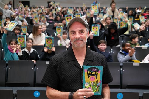 Autor Nils Mohl mit seinem Buch "Die Insel der Schlasocks" in einem Hörsaal der Universität Siegen, im Hintergrund sitzen viele Kinder, die sein Buch in die Höhe halten.
