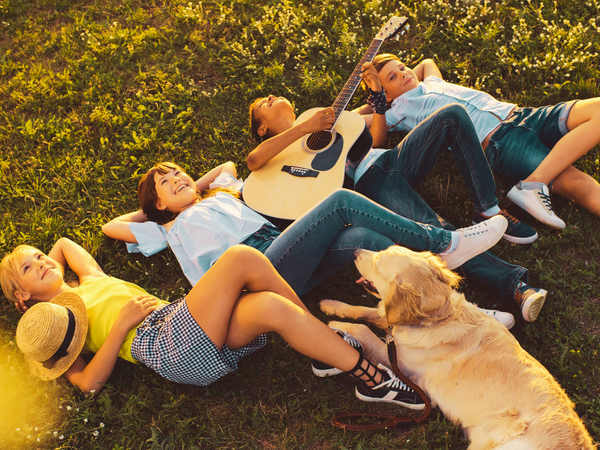 Das Bild zeigt eine Gruppe von vier Jugendlichen, die gemeinsam mit einem Golden Retriever entspannt im Gras liegen. Ein Junge spielt dabei auf einer Akustikgitarre. 